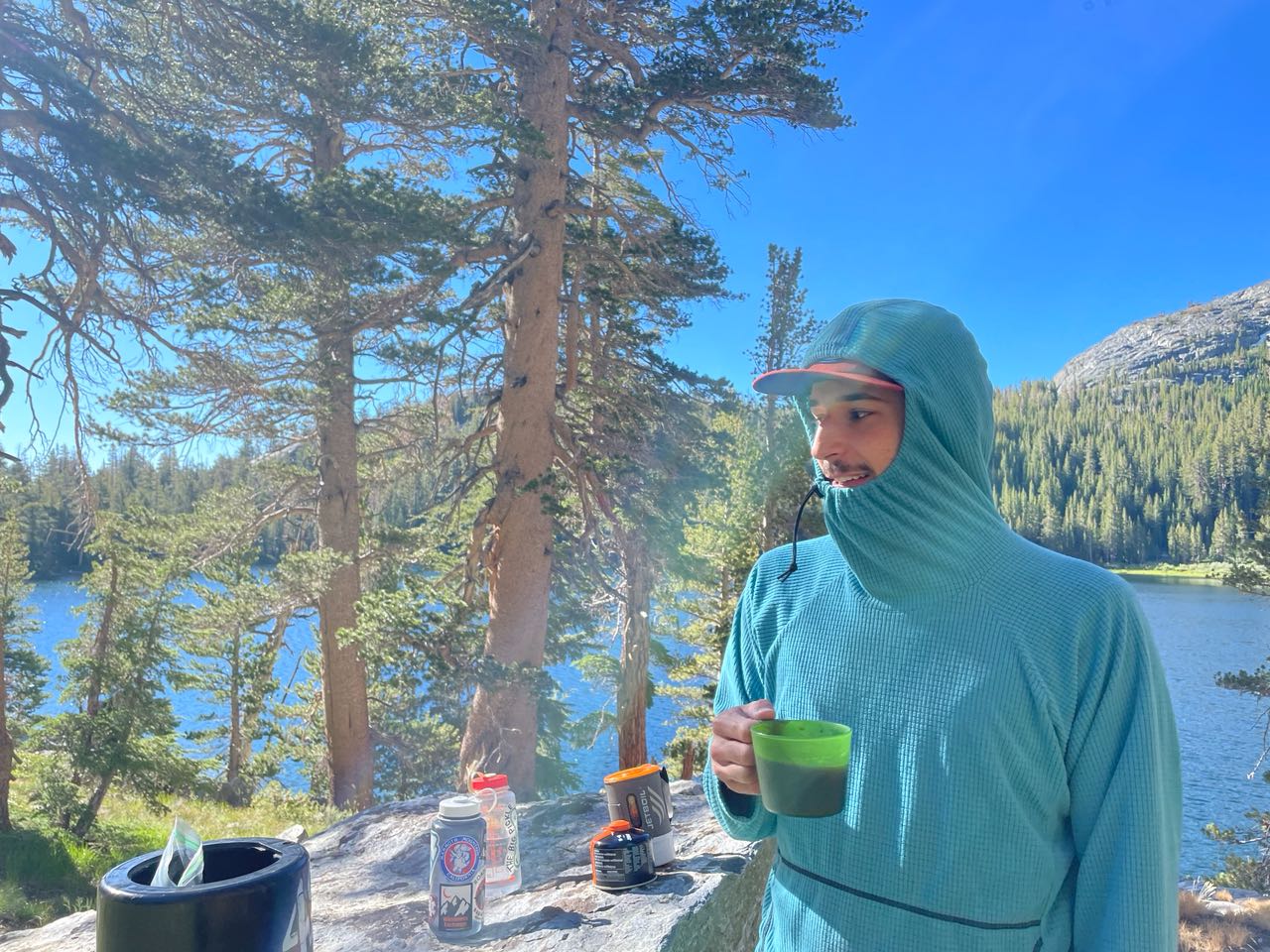 A hooded hiker holding a mug beside an alpine lake ringed by pines